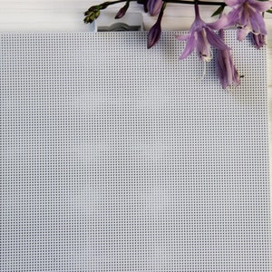 Może przedstawiać: Biały materiał Aida z szpulką jasnoniebieskiej nici do haftu i fioletowym kwiatem na białym drewnianym tle. Szpulka nici ma nadrukowany numer 3754.
