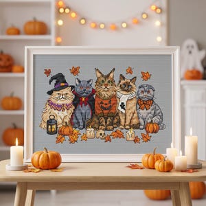 Könnte beinhalten: Ein gerahmtes Kreuzstich-Stickbild mit fünf Katzen in Halloween-Kostümen, darunter ein Hexenhut, ein Umhang und ein Kürbis-Pullover, umgeben von orangefarbenen Herbstblättern, kleinen Kürbissen, Kerzen und einem Totenkopf auf grauem Grund.