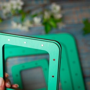Op de afbeelding: Close-up van verschillende vierkante, turquoise houten frames met kleine zilveren en rode accenten. De frames zijn gestapeld, met &eacute;&eacute;n in de hand. De achtergrond is wazig, met groen en een blauw getint oppervlak.