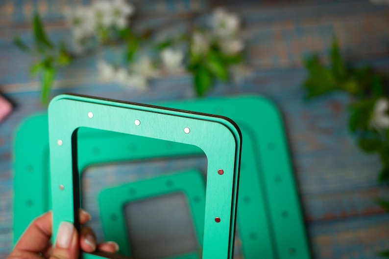 Op de afbeelding: Close-up van verschillende vierkante, turquoise houten frames met kleine zilveren en rode accenten. De frames zijn gestapeld, met &eacute;&eacute;n in de hand. De achtergrond is wazig, met groen en een blauw getint oppervlak.
