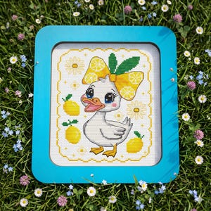 Pode incluir: Imagem de ponto cruz de um pato de desenho animado com um laço de limão amarelo e limões. A obra de arte é emoldurada em uma moldura quadrada azul brilhante. O fundo é branco com detalhes amarelos. O pato tem olhos azuis e pés amarelos.
