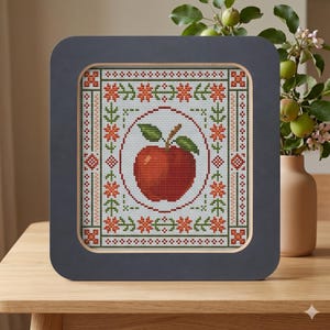 Op de afbeelding: Een ingelijste kruissteek met een rode appel met groene bladeren, omringd door bloemen en geometrische patronen in rood, groen en wit. De donkergrijze lijst heeft een gouden binnenrand. Het kunstwerk wordt op een houten oppervlak weergegeven.