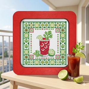 May include: A square red wooden frame containing a cross-stitch pattern of a red cocktail in a glass with a celery stalk and lime slice, surrounded by a decorative border of green lime slices and small red and green geometric shapes.