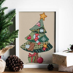 Op de afbeelding: Ingelijste kruissteekkunst van een kerstboom met een gouden ster. De boom is versierd met diverse ornamenten, cadeaus en een rode bal. Het kunstwerk staat in een witte lijst, met een neutrale achtergrond.
