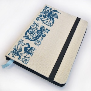 May include: A white canvas notebook with a blue embroidered design of stylised birds and animals. The notebook has a black elastic closure.
