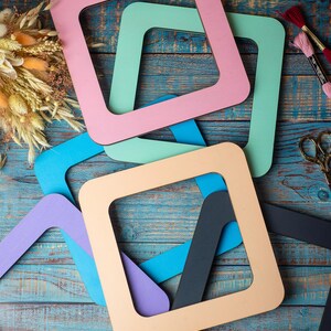 Op de afbeelding: Een verzameling kleurrijke, vierkante houten frames in pastelkleuren roze, groen, blauw en beige, gerangschikt op een verweerd blauw houten oppervlak. Knutselbenodigdheden, waaronder borduurgaren en schaar, zijn ook zichtbaar.