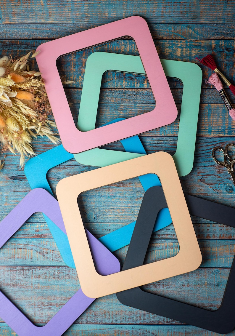 Op de afbeelding: Een verzameling kleurrijke, vierkante houten frames in pastelkleuren roze, groen, blauw en beige, gerangschikt op een verweerd blauw houten oppervlak. Knutselbenodigdheden, waaronder borduurgaren en schaar, zijn ook zichtbaar.