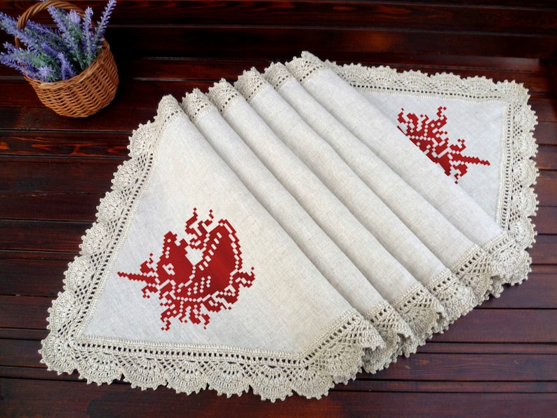 May include: A beige linen table runner with a crocheted border and two red embroidered unicorn designs.