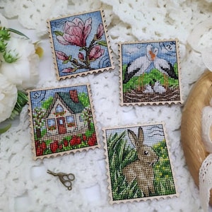 May include: Four cross-stitch art pieces resembling postage stamps. The designs include a pink magnolia flower, a pair of storks with their chicks, a house with a red door, and a brown rabbit in tall grass. Each piece has a wooden frame.
