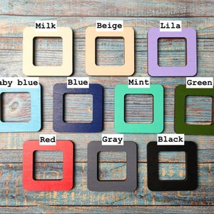 Op de afbeelding: Een verzameling van tien vierkante houten frames in verschillende kleuren, waaronder melk, beige, lila, babyblauw, blauw, mint, groen, rood, grijs en zwart. Elk frame heeft een vierkante uitsparing in het midden.