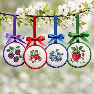 May include: Four round, embroidered ornaments with fruit designs hang from colorful ribbons. The ornaments feature cross-stitch images of blackberries, strawberries, blueberries, and a strawberry. Each ornament has a different colored ribbon and bow.