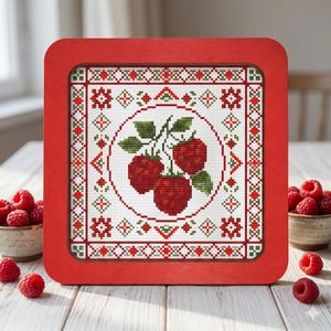 Könnte beinhalten: Ein gerahmtes Kreuzstichmuster mit drei roten Himbeeren auf weißem Grund, umgeben von einem rot-grünen geometrischen Rand. Der Rahmen ist einfarbig rot. Frische Himbeeren befinden sich in kleinen Schalen in der Nähe.