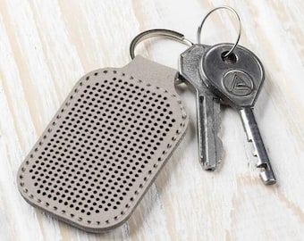 Op de afbeelding: Een grijze leren sleutelhanger met een gestikt ontwerp en een sleutelring. Twee zilveren sleutels zijn aan de sleutelring bevestigd.