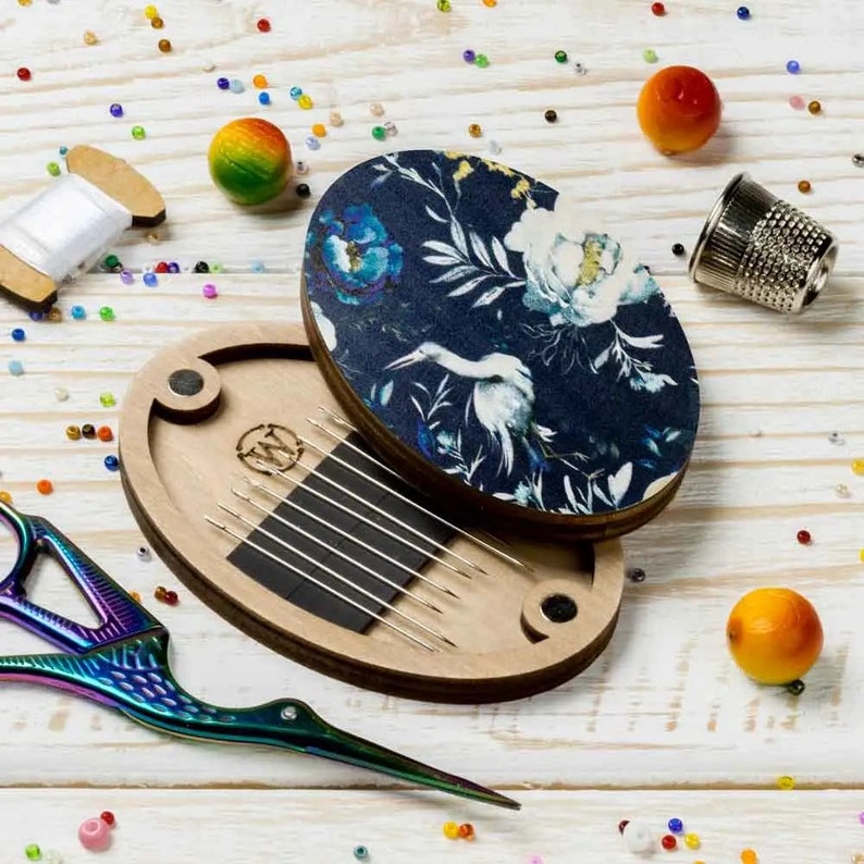 May include: A wooden needle minder with a floral design lid. The lid is blue with white flowers and a white bird. The needle minder has a magnetic closure and holds needles in a slot.