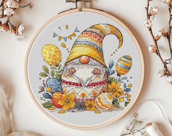 May include: A cross-stitch embroidery hoop with a colorful gnome holding a decorated Easter egg. The gnome is wearing a yellow hat with a blue and red patterned brim. The gnome is surrounded by yellow flowers and green leaves.