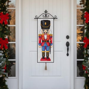 May include: A decorative, rectangular nutcracker banner with a black frame, hanging on a white door. The nutcracker is in a traditional uniform with red, blue, and gold details. A black metal scrollwork hanger and a tassel complete the design.