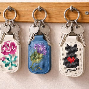 Könnte beinhalten: Drei Schlüsselanhänger mit Schlüsseln, die an einer Holzleiste hängen. Die Schlüsselanhänger haben Kreuzstichmuster: eine rosa Rose, lila Blumen und eine schwarze Katze mit rotem Herz. Die Schlüsselanhänger sind weiß und blau.