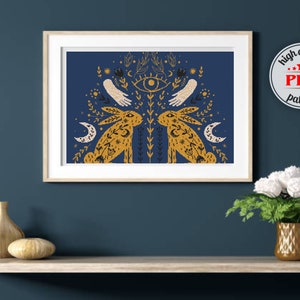 以下が含まれることがあります： 濃い青色の背景に、スタイリッシュなウサギが2匹と、第三の目と花柄のモチーフが描かれたクロスステッチパターン。パターンは額装され、2つの装飾的な花瓶のある棚に飾られています。