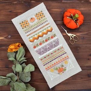 May include: A beige linen fabric with a variety of embroidered designs in orange, green, white, and purple. The designs include pumpkins, leaves, vines, and geometric patterns.