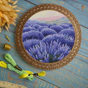 May include: A cross-stitch embroidery of a lavender field in a round wooden frame. The frame has a decorative border. Green and yellow embroidery floss, a thimble, and dried flowers are arranged around the framed artwork.