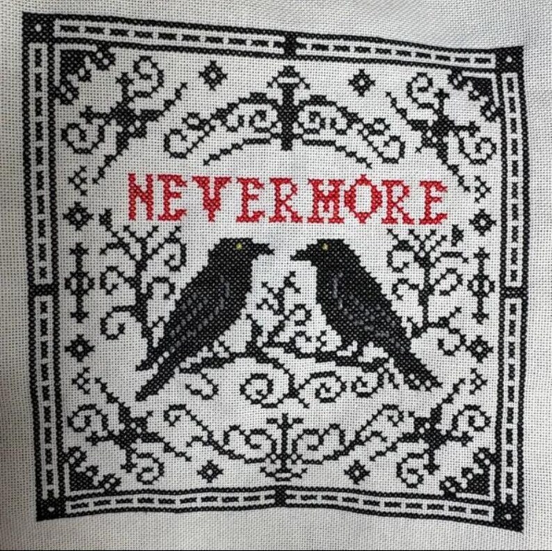 May include: Cross-stitch embroidery on white fabric featuring two black crows perched on a branch, with the red word "NEVERMORE" above them. The design is framed by a black border with decorative elements.