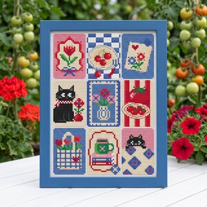 May include: A framed cross-stitch sampler featuring a grid of nine distinct illustrations, including a tulip, cherries, a black cat, a vase of flowers, a plate of food, a tote bag, a mug on books, and a cat peeking over a blue patterned quilt.