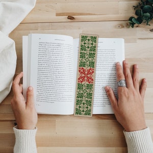 May include: A close-up of a green and red embroidered bookmark with a beige background. The bookmark is made of burlap and has a simple, geometric design.