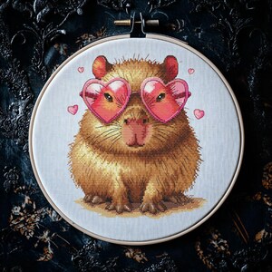 May include: A brown and white capybara wearing pink heart-shaped sunglasses. The capybara is sitting on a white background with pink hearts scattered around it.