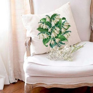 May include: A white square throw pillow with an embroidered floral design featuring white flowers and green leaves. The pillow rests on a white chair, with a small bouquet of white flowers in front of it. The chair has a light wood frame.