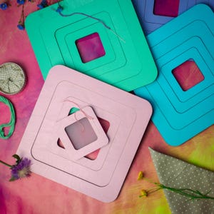 Può includere: Una collezione di telai da ricamo quadrati colorati, nei toni del rosa, verde acqua e blu. Ogni telaio ha un'apertura quadrata più piccola al centro, con tessuto a rete visibile. Un ago e filo sono posizionati sui telai, suggerendo un progetto artigianale.