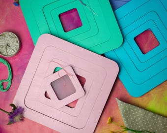 May include: A collection of square wooden embroidery frames in pink, teal, and blue, each featuring concentric square cutouts. A small pink frame sits atop a larger pink one, with thin embroidery thread and needles attached to the surfaces.