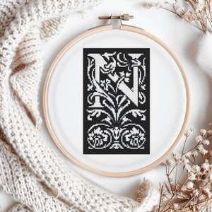 May include: A black and white cross-stitch design of the letter "N" in a wooden embroidery hoop. The letter is in a decorative, floral style. The hoop is on a white surface, with a cream-colored knitted item and dried flowers in the background.