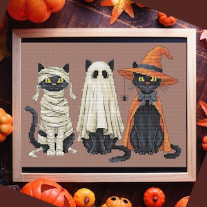 Può includere: Ricamo a punto croce con tre gatti neri vestiti per Halloween. Uno è una mummia, un altro un fantasma e il terzo una strega con cappello e mantello arancione. L'opera è incorniciata in una cornice di legno.
