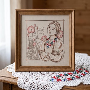 May include: A framed cross-stitch embroidery featuring a portrait of a person with arms crossed, wearing a patterned collar, surrounded by line-art poppies in shades of pink and brown on a light fabric background, set in a light brown wooden frame.
