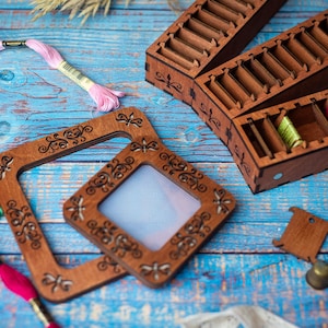 Op de afbeelding: Twee houten borduurringen met ingewikkelde bloemmotieven. De grotere ring is leeg, terwijl de kleinere ring een stuk wit stof bevat. De ringen liggen op een blauw houten oppervlak met spoelen borduurgaren.