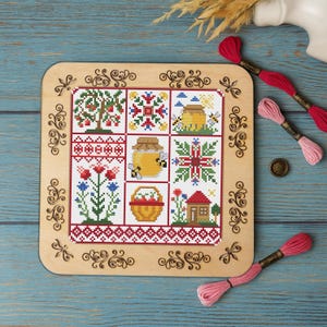 Puede incluir: Un muestrario de punto de cruz con nueve cuadrados con diseños de arte popular, incluyendo árboles, flores y tarros de miel con abejas. El muestrario está enmarcado en un marco de madera con volutas decorativas. Hilos de bordar rojos, rosas y blancos son visibles.