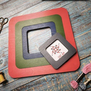 Op de afbeelding: Een verzameling kruissteekbenodigdheden. De afbeelding toont een set rode, groene en blauwe vierkante frames, een kleine kruissteek met het woord "DREAM", een schaar, garenklosjes en borduurgaren. De items zijn geplaatst op een blauw houten oppervlak.