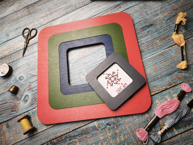 Op de afbeelding: Een verzameling kruissteekbenodigdheden. De afbeelding toont een set rode, groene en blauwe vierkante frames, een kleine kruissteek met het woord "DREAM", een schaar, garenklosjes en borduurgaren. De items zijn geplaatst op een blauw houten oppervlak.