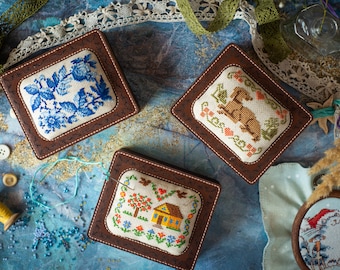 Op de afbeelding: Drie kleine, vierkante, bruine lederen doosjes met geborduurde ontwerpen. De doosjes staan op een blauw-witte, getextureerde ondergrond. De ontwerpen zijn een blauw bloemenpatroon, een bruine hond en een geel huis met een boom.