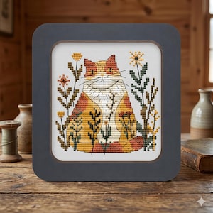 Peut inclure: Une broderie au point de croix dans un cadre carré représentant un chat stylisé orange et blanc assis parmi des fleurs sauvages vert foncé et jaunes. Le chat a un poitrail blanc et des taches orange, avec des traits noirs pour le visage.