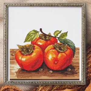 May include: Cross-stitch artwork of three ripe persimmons with green leaves. The persimmons are a vibrant orange color, and the leaves are a mix of green and brown. The artwork is framed in a silver-colored frame.