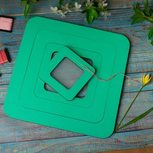 Op de afbeelding: Een teal groene vierkante borduurring set met een naald en draad, omringd door kleurrijke spoelen garen. De items zijn gerangschikt op een verweerd blauw houten oppervlak, met bloemaccenten.