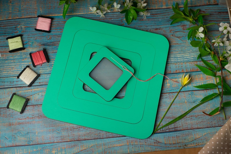 Op de afbeelding: Een teal groene vierkante borduurring set met een naald en draad, omringd door kleurrijke spoelen garen. De items zijn gerangschikt op een verweerd blauw houten oppervlak, met bloemaccenten.
