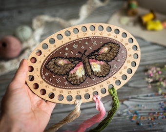 May include: An oval wooden embroidery floss organizer featuring a brown leather-like center panel with a cross-stitched moth design. The wooden frame has thirty-two numbered holes along the perimeter, with three strands of thread attached.
