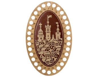 May include: An oval wooden embroidery floss organizer featuring a dark brown leather center inlay with a cream-colored cross-stitch pattern of a castle and town. The wooden frame has thirty-two circular holes and numbers 1 through 32.
