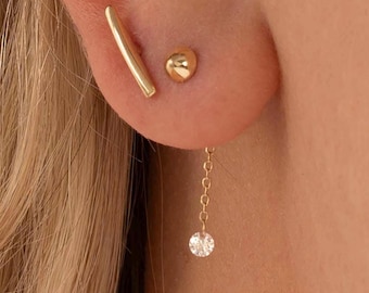 May include: Gold earrings with a small diamond dangle and a thin bar stud. The earrings are worn in the earlobe and helix.
