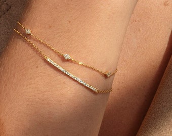 May include: Two delicate gold bracelets with small, sparkling gemstones. One bracelet features a line of gems, while the other has scattered stones. The bracelets are worn on a wrist, against a neutral background.