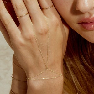 Könnte beinhalten: Ein goldenes Kettenarmband mit zwei zarten Ketten, die sich vom Handgelenk zu den Fingern erstrecken. Das Armband wird am linken Handgelenk getragen.