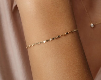 May include: A delicate gold bracelet with small, flat, rectangular links is worn on an arm. A matching gold necklace with a small pendant is also visible. The background is a soft, light pink.