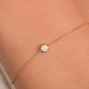 May include: A fine gold bracelet with a single, round, clear gemstone. The bracelet has a delicate chain design, with the gemstone set in a bezel. The bracelet is shown on a wrist, against a neutral background.